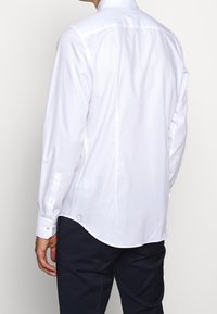 Bílá košile s dlouhým rukávem, zakřiveným lemem, zapínáním na knoflíky na manžetách a hladkou texturou, zobrazena zezadu, v kombinaci s tmavě modrými kalhotami.