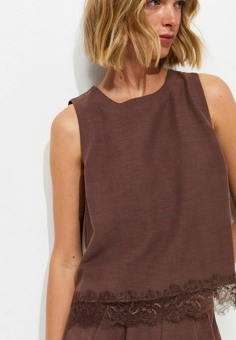 Woman with short blonde hair wearing a sleeveless brown top with lace trim at the hem, looking to the side against a plain background.