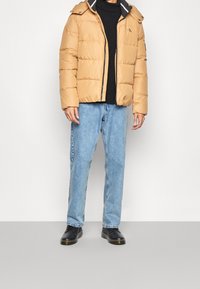 Beige pufferjakke med vandret syninger, lynlås lukning og hætte, parret med lyseblå jeans og sorte sko. Sort sweater under.