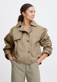 Giacca beige doppiopetto con ampio colletto, vestibilità oversize e dettagli in bottoni. Presenta un tessuto liscio e leggero e maniche arrotolate.