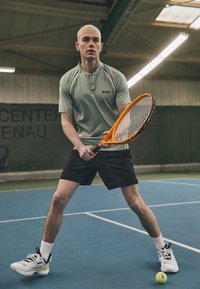 Zielona koszulka polo z czarnymi akcentami, czarne szorty i białe trampki. Trzyma pomarańczową rakietę tenisową, gotowy do gry na niebieskim korcie.