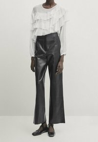 Blusa bianca con rouches e maniche lunghe abbinata a pantaloni larghi in pelle nera a vita alta e scarpe nere senza lacci.