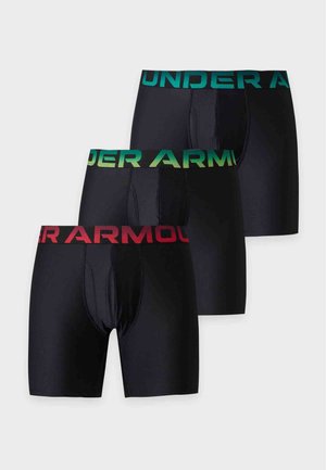 Muskulovitý muž stojaci bosý v čiernych boxerkách s červeným pásom a textom "ARMOUR" na jednoduchom svetlom pozadí.