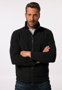 Chaqueta negra con cremallera y cuello alto, de tejido texturizado, con bolsillos laterales y un logo en la manga izquierda, llevada sobre una camisa blanca.