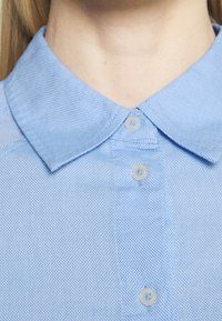 Chemise bleu clair à manches courtes avec un tissu texturé. Présente un col classique et trois boutons au niveau du cou.