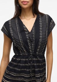 A woman smiles, wearing a dark dress with a V-neck and cap sleeves, featuring a patterned, cinched waist design.