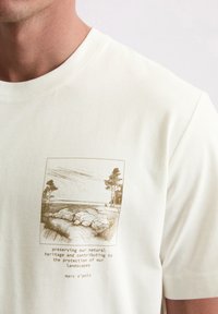 Prim-plan al unei cămăși albe cu un desen maro al unei linii de coastă stâncoase și copaci, având text despre conservarea patrimoniului natural.
