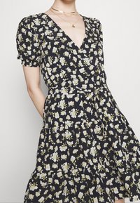 Robe portefeuille noire avec un décolleté en V, des manches courtes bouffantes et une jupe à volants. Ornée de petits motifs floraux blancs et jaunes.