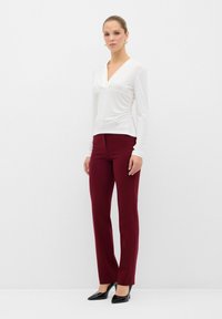 Maglietta bianca a maniche lunghe con scollo a V e dettagli arricciati; abbinata a pantaloni bordeaux su misura; décolleté nere a punta completano l'outfit.