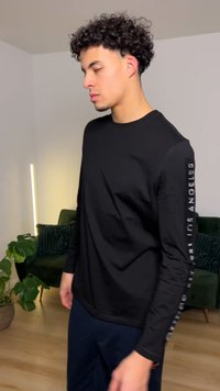 Crna majica dugih rukava od mekane tkanine, s tekstom na lijevoj rukavini, okruglim izrezom i opuštenim krojem, uparena s mornarsko plavim hlačama.