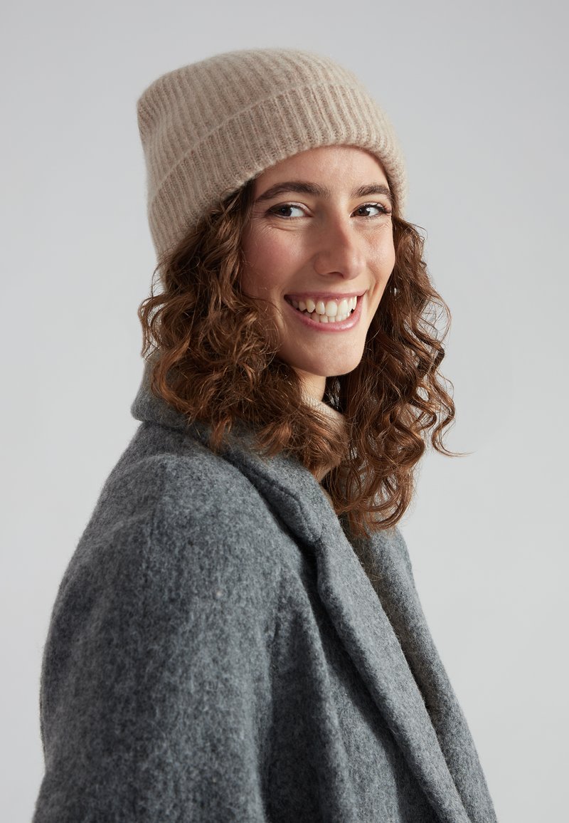 Beanie a coste beige, realizzato in morbida lana, indossato da una persona in un cappotto di lana grigio, con capelli ricci e un sorriso caloroso.