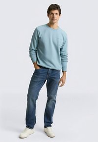 Lichtblauwe trui met geribbelde manchetten en boord, gecombineerd met donkerblauwe spijkerbroek en witte sneakers. Eenvoudig ontwerp, zonder patronen of zichtbare logo's.