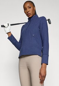 Pullover de golf de color azul marino con cremallera de media longitud, hecho de un tejido texturizado, que presenta un sutil bolsillo lateral y un ajuste relajado, combinado con pantalones ajustados de color beige claro.
