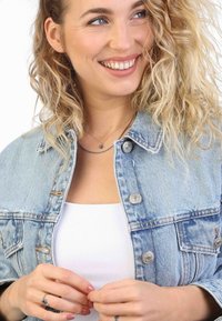 Hellblaues Denim-Jacket mit silbernen Knöpfen, über einem weißen Oberteil getragen. Das Model hat lockiges Haar und trägt geschichtete silberne Halsketten.