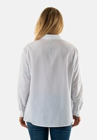Camisa blanca de manga larga con dobladillo recto, costura de yugo en la parte posterior y puños abotonados. La tela parece ligera y suave.