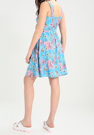 Femme portant une robe bleue sans manches avec un imprimé floral rose et blanc, debout de dos, chaussée de sandales à plateforme blanches.