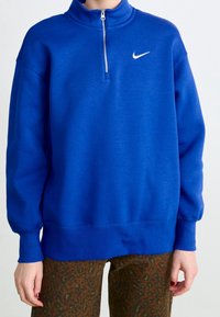 Modrá mikina Nike se zipem na vysokém límci, dlouhými rukávy a žebrovaným lemem. Na hrudi je malé bílé logo.
