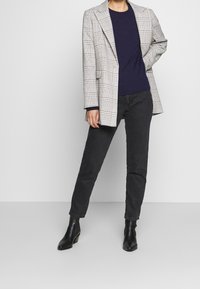 Grau kariert Blazer mit strukturierten Schultern, kombiniert mit einem navyblauen Pullover und schwarzen Jeans. Schwarze Stiefeletten mit Absatz vervollständigen das Outfit.