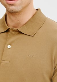 Brązowa koszulka polo z teksturowanej tkaniny, z dwoma guzikami przy kołnierzyku i wytłoczonym logo na piersi. Widoczne krótkie rękawy.