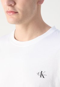 Camiseta blanca de algodón con cuello redondo, que presenta un pequeño logo negro de Calvin Klein Jeans en el pecho, textura suave, diseño casual.