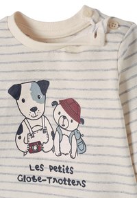 Gestreiftes Creme-Oberteil mit illustrierten Hund- und Bärenfiguren, Knöpfen an der Schulter und dem Text "Les Petits Globe-Trotters" darunter.