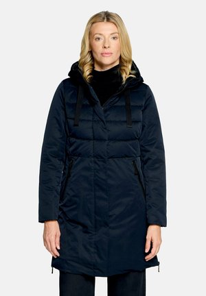 Femme aux cheveux blonds bouclés portant un long manteau d'hiver matelassé bleu foncé avec capuche, debout devant un fond clair uni.