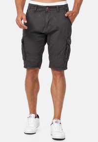 Homme portant un short cargo noir avec des poches latérales et des baskets blanches Reebok, debout devant un fond clair uni.