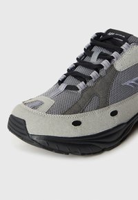 Pelēka sporta apava ar teksturētu tīkliņa augšdaļu, zamšādas akcentiem, melnām šņorēm un gumijas zolīti ar ventilācijas caurumiem gaisa caurlaidībai.