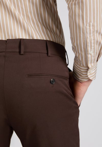 Homme portant un pantalon marron avec une poche arrière à bouton et une chemise à manches longues beige à rayures, main posée sur la hanche.