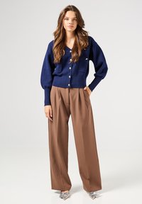 Cárdigan de punto azul marino con escote en V, bolsillos frontales y botones plateados, combinado con pantalones de talle alto marrones de pierna ancha y zapatos plateados de punta.