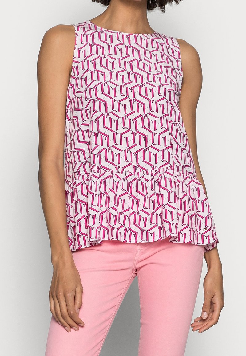 Top peplum senza maniche in tessuto bianco con un motivo geometrico rosa. Texture morbida con orlo svasato, abbinato a pantaloni rosa.