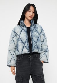 Jonge vrouw met lang zwart haar, gekleed in een blauwe gewatteerde pufferjack en zwarte cargobroek, staand met een hand in haar zak.
