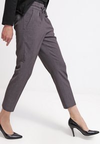 Pantalones gris ajustados con cintura elástica, bolsillos laterales y pierna tapered, combinados con tacones de punta negros. Textura de tela suave.
