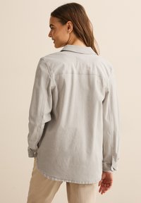 Camicia a maniche lunghe con colletto di colore grigio chiaro con strisce bianche verticali. Presenta un orlo curvo e polsini con bottoni. Tessuto morbido e texturizzato.