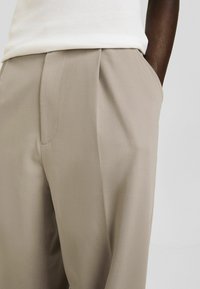 Pantalones beiges ajustados con una parte delantera plana, bolsillos laterales y una textura suave. La tela cae con elegancia con pliegues sutiles para un detalle adicional.
