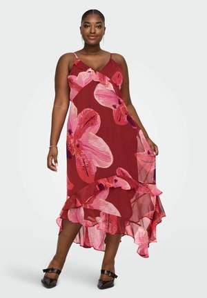 Kvinne iført en rød ermeløs kjole med stort rosa blomstermønster og rysjet asymmetrisk kant, kombinert med svarte spisse sko.