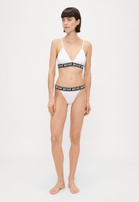 Bijeli bikini s trokutastim braletom i minimalno pokrivenim donjim dijelom. Crni elastični pojas sadrži "justcavalli" logo. Glatka tekstura tkanine.