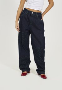 Dark blue denim jeans with a loose fit, green stitching, and large graphic designs on the legs. Styled with red block-heeled shoes.