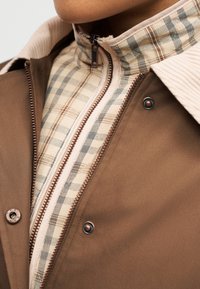 Κοντινό πλάνο από στρωμένες μπεζ και καφέ ζακέτες με εμφανείς φερμουάρ και κουμπιά πάνας στο πάνω μέρος του στήθους και τον λαιμό ενός ατόμου.