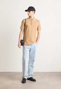 Odtisnjen polo majica v temno bež barvi z belimi detajli, svetlo modri kavbojki in črnimi čevlji. Model nosi črn čez ramo nahrbtnik in ima na glavi črno kapo.
