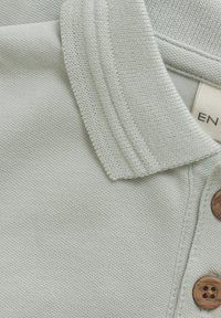 Ljusgrön polo med en texturerad tygkvalitet, en ribbad krage och tre bruna knappar längs placket. Varumärkesetikett synlig.