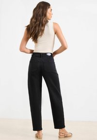 Mujer de pie con las manos en las caderas, vistiendo una blusa sin mangas beige, pantalones vaqueros negros de cintura alta y zapatos planos beige, de espaldas.
