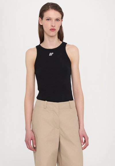 Schwarzes ärmelloses Top mit Rundhalsausschnitt und weißem Logo auf der Brust, kombiniert mit hellbeigen, hochgeschnittenen Hosen. Glatte Stofftextur.