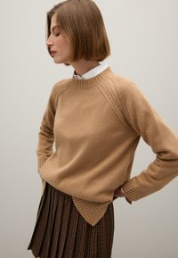 Beige gestrickter Pullover mit hohem Rippen-Ausschnitt und Raglanärmeln. Tragen über einem dunkel karierten Faltenrock mit dezenten roten Akzenten.