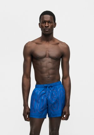 Άνδρας χωρίς πουκάμισο με σκούρο δέρμα φορά μπλε μαγιό με έντονα γράμματα με περίγραμμα, στέκεται μπροστά σε απλό ανοιχτό γκρι φόντο.