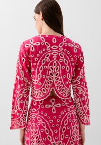 Haut fuchsia brodé avec des manches longues et un dos ouvert, présentant des motifs floraux et de feuilles blancs complexes sur un tissu texturé.