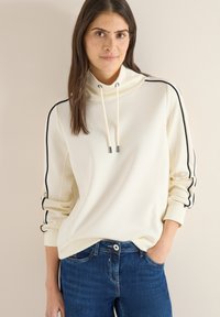 Lichtbeige sweatshirt met hoge kraag, trekkoord en zwarte strepen aan de zijkanten. Zachte textuur, comfortabele pasvorm, gecombineerd met donkerblauwe jeans.