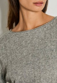 Frau trägt einen hellgrauen Kabelstrickpullover mit weitem Ausschnitt, der einen Teil des Halses und der Lippen im neutralen Licht zeigt.