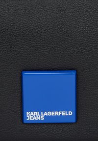 Juodas tekstūruotas odos paviršius su mėlynu kvadratiniu logotipo plokštelė, ant kurios baltais raidėmis užrašyta "KARL LAGERFELD JEANS".