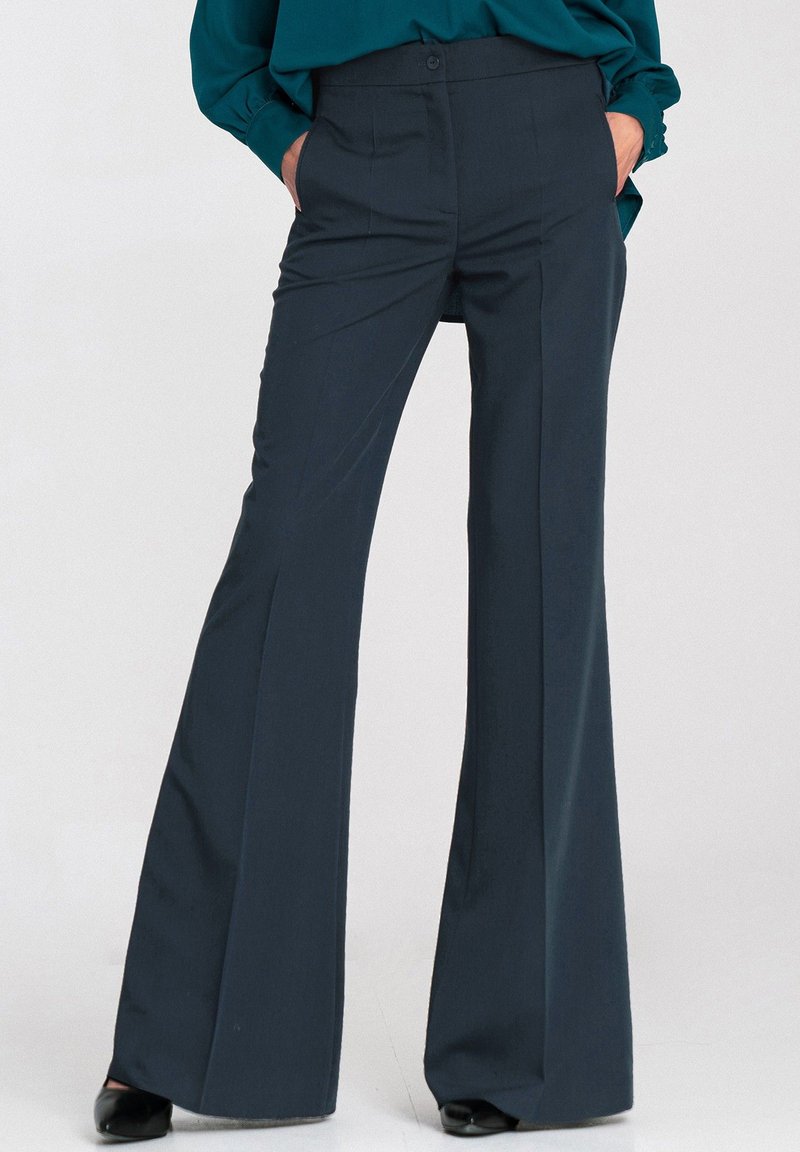 Kobieta w ciemnogranatowych, wysokich spodniach z szerokimi nogawkami i frontowym zagięciem, z dłońmi w kieszeniach, w połączeniu z czarnymi butami i turkusową bluzką.